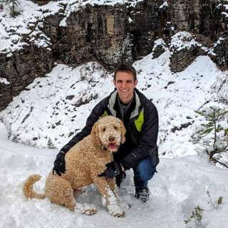 Chad March kneeling in the snow next to Pivo, a curly-coated dog